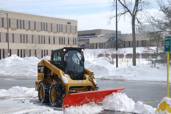 A skid steer with a snow plow clearing snow near a commercial building, highlighting the commercial landscaping bid process.