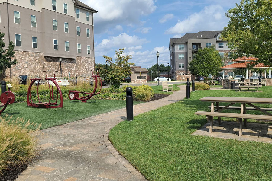 Corporate campus landscape design featuring a walking path, outdoor seating, greenery, and exercise equipment near modern offices.