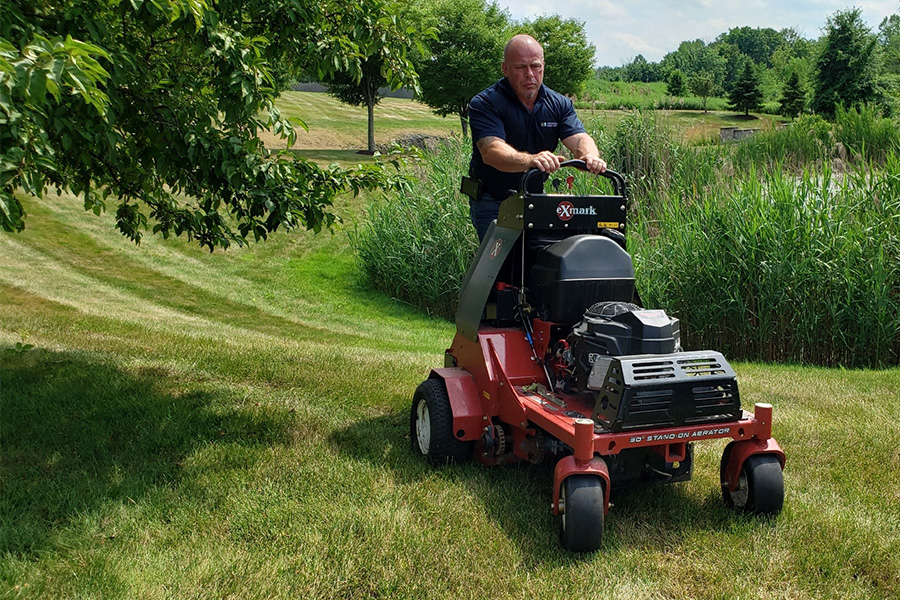 Commercial landscape maintenance worker operating a lawn aerator machine on a well-kept grassy area with trees in the background