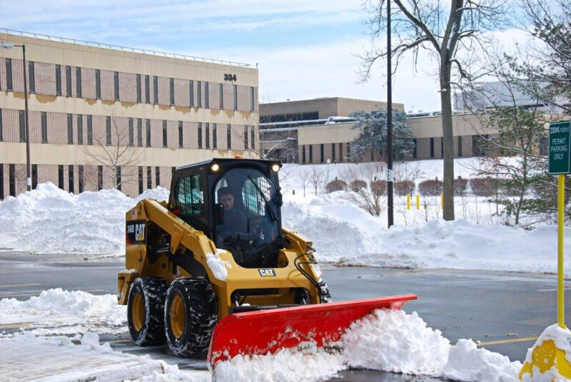 Skid-steer loader with snow plow clearing a commercial lot, illustrating commercial landscaping safety ideas for winter maintenance