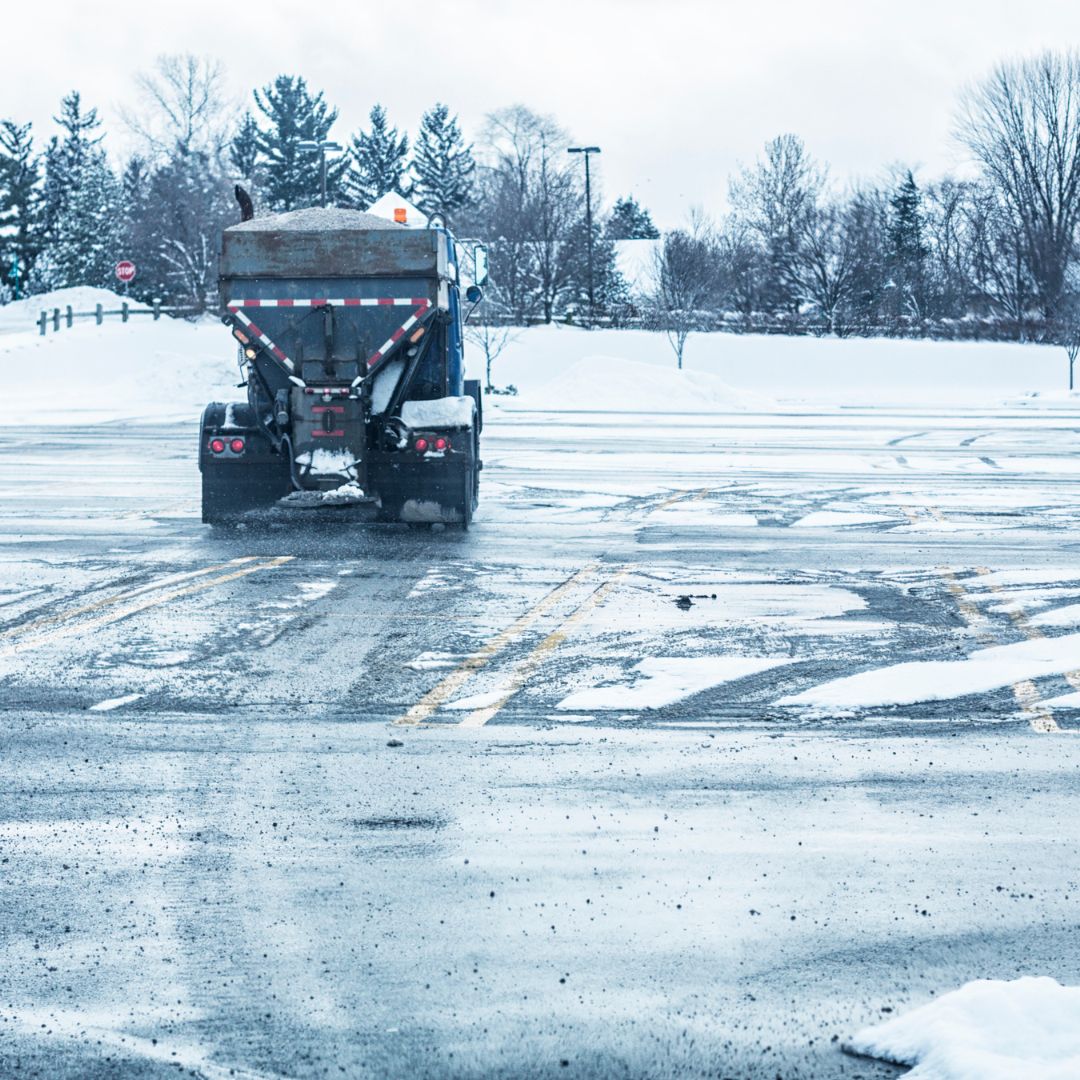 A snow truck with plow in a commercial lot applying salt to a parking lot