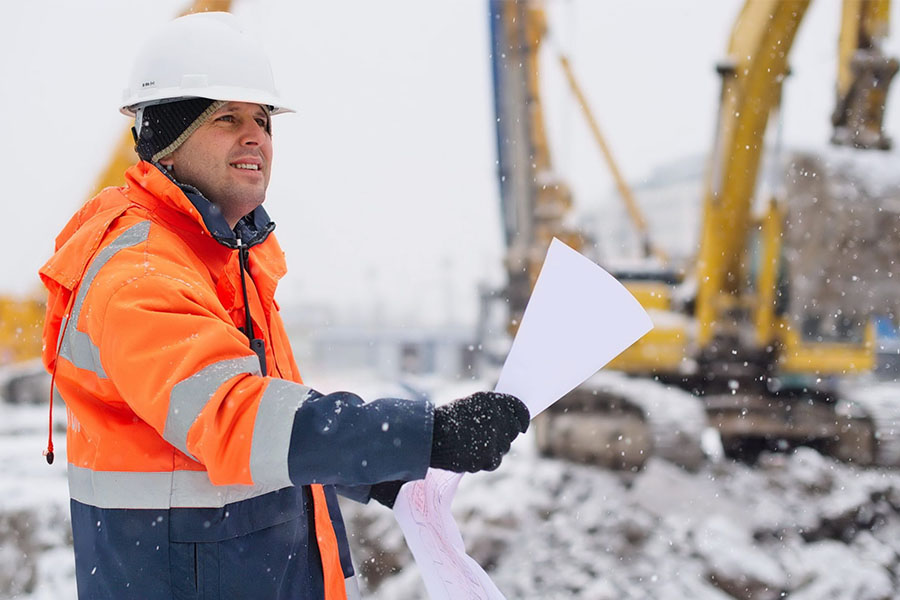 Worker preparing commercial property for severe weather on a snowy construction site, wearing safety gear and holding documents